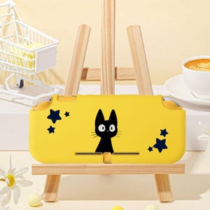 May include: A yellow handheld gaming console case with a black cat design and dark blue star details. The case is presented on a small wooden easel. The background includes a miniature shopping trolley and a cup of coffee.