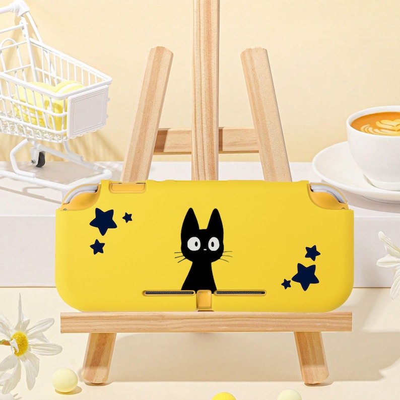 May include: A yellow handheld gaming console case featuring a black cat graphic and dark blue star accents. The case is displayed on a small wooden easel. The background includes a miniature shopping cart and a cup of coffee.