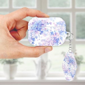 Op de afbeelding: Witte AirPod-hoes met een bloemenontwerp in blauwe en paarse tinten. De hoes heeft een bijpassende sleutelhanger met hetzelfde bloemenpatroon. De hoes wordt voor een raam gehouden.