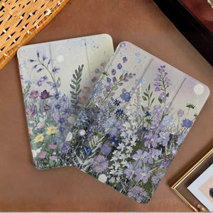 Puede incluir: Dos fundas para tabletas con un diseño floral. Las fundas presentan una ilustración al estilo acuarela de flores silvestres y vegetación moradas, azules y amarillas sobre un fondo claro. Las fundas son rectangulares con esquinas redondeadas.