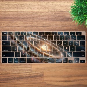 May include: A keyboard skin featuring a galaxy design. The keys are covered with a cosmic pattern of swirling nebulae, planets, and stars in shades of brown, blue, and gold. The keyboard is set against a wooden surface.