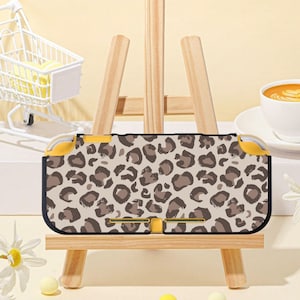 Puede incluir: Una funda para Nintendo Switch Lite con estampado de leopardo en beige y marrón. La funda tiene detalles amarillos y se muestra en un pequeño caballete de madera. La funda mide aproximadamente 19 cm de largo.