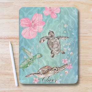 May include: Tablet case featuring a watercolor design of sea turtles swimming among pink hibiscus flowers and bubbles. The background is a light blue with a wavy pattern. The name "Oliver" is written in cursive.