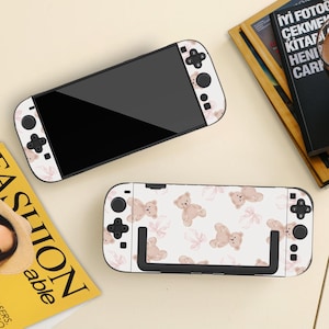 May include: Two white Nintendo Switch consoles featuring a teddy bear and pink bow design. The consoles are on a light beige surface, with a magazine and books in the background. The controllers have black buttons.