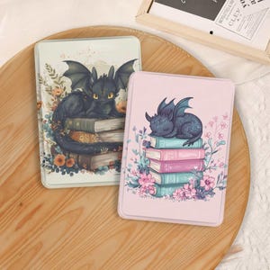 May include: Two rectangular e-reader cases featuring dragon illustrations. The left case shows a black dragon perched on a stack of books surrounded by orange flowers. The right case is pink and depicts a small blue dragon sleeping on colorful books.