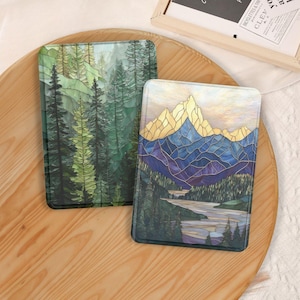 May include: Two rectangular protective cases featuring stained glass style artwork. One case displays a dense forest of green pine trees. The other case depicts a mountain range with golden peaks, blue slopes, and a river flowing through a forest.