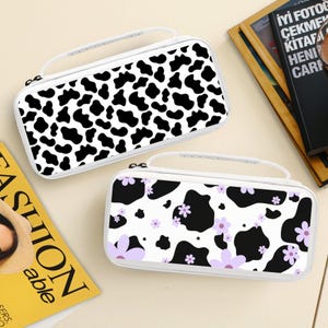 May include: Two white carrying cases with black cow print patterns. One case has a classic cow print, while the other features a cow print with lavender flowers. Both cases have white handles and zippers.