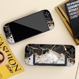 May include: Two gaming console cases with a black and gold marble pattern. The cases have a black screen and black buttons. The marble design features white and gold veining. The cases are on a light beige surface.