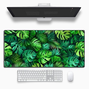 May include: A large rectangular desk mat with a vibrant green tropical leaf pattern. The mat is positioned beneath a computer monitor, a white keyboard, and a white mouse. The design includes various shades of green leaves.