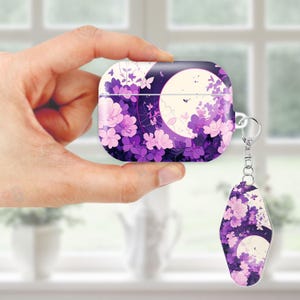 May include: A purple and white AirPod case with a matching keychain. The case and keychain feature a floral design with a full moon and butterflies. The case is held in a hand.