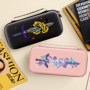May include: Two rectangular game console cases. One is black with a sword and rose design. The other is pink with a sword and floral design. Both cases have a zipper closure and a handle.