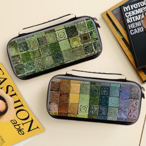 Puede incluir: Dos estuches rectangulares con marco negro y asa blanca. Los estuches presentan un diseño de mosaico con varios colores y patrones. Un estuche tiene azulejos verdes y el otro tiene un degradado de colores. Los estuches están sobre una superficie clara.