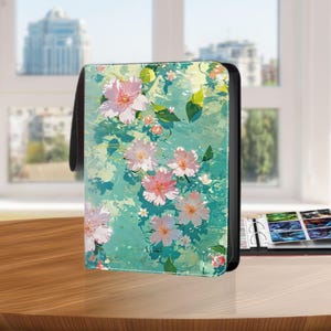 May include: A black binder with a floral design featuring pink flowers and green leaves on a teal background. The binder has a black handle and is sitting on a wooden surface.