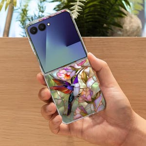 May include: A clear smartphone case featuring a colorful hummingbird design. The hummingbird has purple, red, and blue feathers, surrounded by pink and green floral elements. The phone is open, displaying a blue and purple screen.