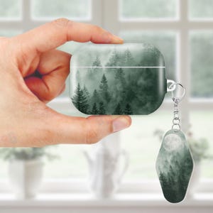 May include: A forest-themed AirPod case with a matching keychain. The case and keychain feature a misty forest scene in shades of green and gray. The keychain has a full moon design. The case is held in a hand.