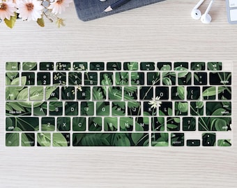 May include: A keyboard cover featuring a green and white tropical leaf design. The cover is placed on a wooden desk, with a phone, pencil, and headphones visible in the background. The keyboard keys are clearly visible.