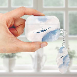 May include: White AirPod case and matching keychain featuring a watercolor design of a blue airplane flying through clouds. The case has a silver keyring attachment. The design is a light blue and white color scheme.