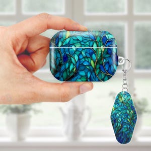May include: A teal and blue AirPod case with a matching keychain. The case and keychain feature a leaf-like design with gold accents. The case is held in a hand, showcasing the intricate pattern and color variations.