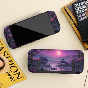 Puede incluir: Dos consolas de juegos portátiles con un diseño morado y negro. Una consola tiene una pantalla negra y detalles morados. La otra consola tiene un paisaje con luna llena, árboles y una pagoda.