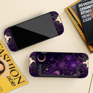 Pode incluir: Um console de jogos portátil preto com um design floral em tons de roxo, creme e dourado. O design inclui flores, uma lua crescente e borboletas. O dispositivo é mostrado com uma capa protetora correspondente.