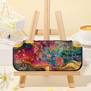 Könnte beinhalten: Eine gemusterte Switch Lite-Hülle mit einem floralen Design in Rosa, Gelb und Türkis. Die Hülle wird auf einer kleinen Holzstaffelei präsentiert. Ein Miniatur-Einkaufswagen und eine Tasse Kaffee befinden sich im Hintergrund.