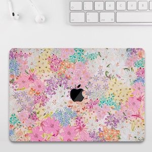 Puede incluir: Una funda para portátil con un vibrante diseño floral en tonos rosa, morado, amarillo y azul. El diseño es un collage de varios tipos de flores, con un logotipo de Apple negro en el centro. La funda está sobre una superficie blanca, con un teclado y auriculares visibles.