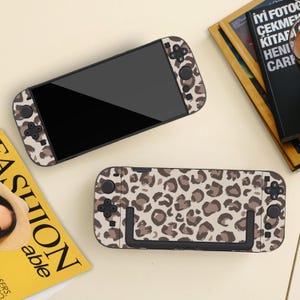 May include: Two gaming console controllers with a leopard print design. The controllers have black screens and buttons, and a beige and brown spotted pattern. They are on a light beige surface, with a magazine and book in the background.