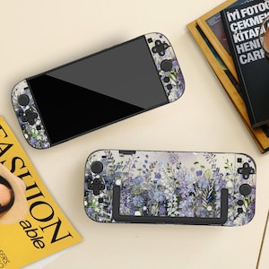 May include: Two Nintendo Switch consoles with floral patterns. The top console is black with floral detailing on the sides. The bottom console features a floral design with purple and blue flowers. The consoles are on a light surface.