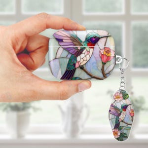 May include: A white AirPod case and matching keychain featuring a stained-glass hummingbird design. The hummingbird has purple, green, and red details, perched on a branch with pink flowers. The case is held in a hand.