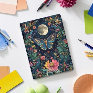 May include: A notebook with a dark blue cover featuring a butterfly, full moon, and floral design. The butterfly is blue and gold, surrounded by colorful flowers. The name "Oliver" is written on the bottom.