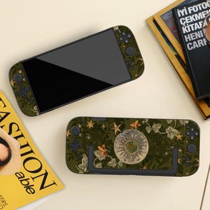 May include: A handheld gaming console and its dock, both featuring a dark green skin with a botanical pattern of ivy leaves, small orange moths, and gold stars, centered by a circular celestial illustration of a sun and moon design.