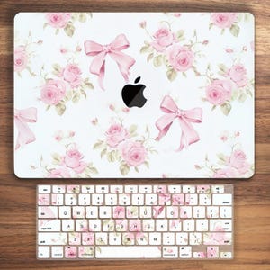 May include: A white laptop computer with a floral design featuring pink roses and bows. The laptop has a black Apple logo. The keyboard is also white with a floral design.