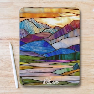Pode incluir: Uma capa para tablet com um design de vitral de uma paisagem montanhosa com um pôr do sol. A capa tem um design colorido com tons de azul, roxo, laranja e verde. O nome "Oliver" está escrito em itálico na parte inferior.