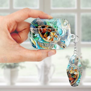 May include: An AirPod case and keyring with a stained-glass effect sea turtle design. The case is rectangular with a curved top, and the keyring is oval. The turtle has a brown, orange, and tan shell with blue and green accents, creating a vibrant look.