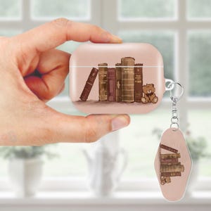 May include: A blush-colored AirPod case and keychain featuring a book-themed design. The case and keychain depict a stack of vintage books and a teddy bear. The case has a silver-colored metal ring for the keychain.
