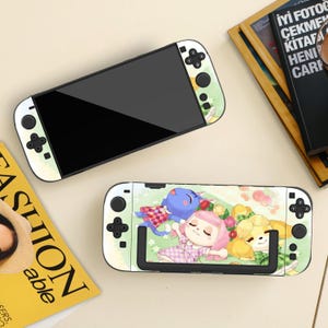 May include: Two Nintendo Switch consoles with custom designs. One console is black with white and green accents. The other console has a colorful design featuring cartoon characters. The consoles are on a light-colored surface.