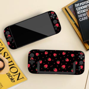 May include: Black handheld gaming console with a strawberry design. The console features a black screen and red strawberries with green leaves on a black backdrop. The pattern is repeated on the controllers.