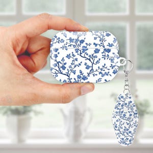 May include: White and blue floral patterned AirPod case with a matching keychain. The case is rectangular with rounded edges and a silver keyring. The floral design features blue flowers and branches on a white background.