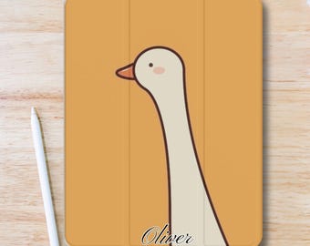 Puede incluir: Una funda para tableta de color amarillo mostaza con una ilustración de un ganso de dibujos animados. El ganso tiene un cuello largo y un pico naranja. El nombre "Oliver" está escrito en cursiva en la parte inferior. Un lápiz óptico blanco está a la izquierda.