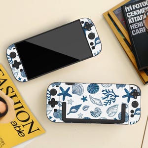 Può includere: Due skin per console di gioco con un motivo a conchiglie blu su sfondo bianco. Il design include stelle marine, conchiglie e coralli. La console ha uno schermo nero e pulsanti neri. Il motivo si ripete sui controller.