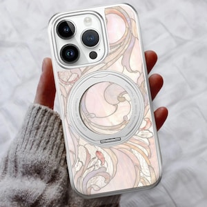 May include: A clear phone case featuring a floral pattern in pink, beige, and purple hues. It has a silver magnetic ring and a transparent border. The phone's camera lenses are visible.