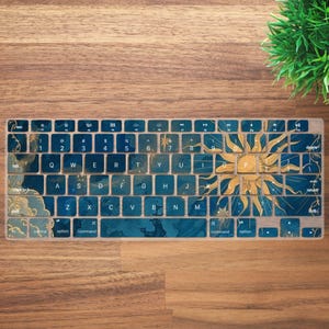 Può includere: Una cover per tastiera con un design a sole blu e oro. La cover ha uno sfondo blu con accenti dorati e una grande grafica solare sul lato destro. I tasti sono visibili con lettere bianche.