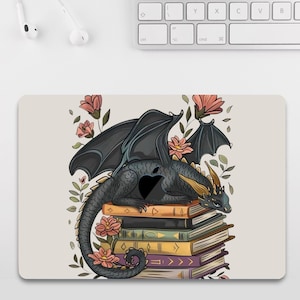 May include: A rectangular mouse mat featuring a black dragon with a heart-shaped cutout, resting on a stack of books. The dragon has large wings and is surrounded by pink flowers and green leaves. The books are various colours.