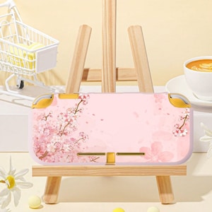 Pode incluir: Uma capa rosa para Nintendo Switch Lite com um design de flor de cerejeira. A capa tem detalhes amarelos nas laterais. É exibida em um pequeno cavalete de madeira, com cerca de 17 cm de comprimento.