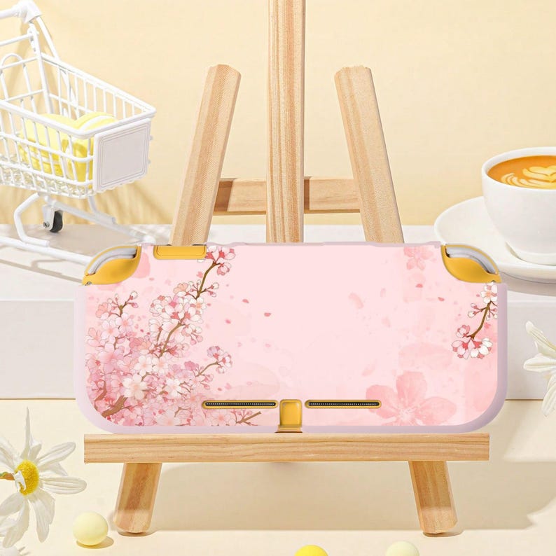 May include: A pink Nintendo Switch Lite case featuring a cherry blossom design. The case has yellow accents on the top and sides. The case is displayed on a small wooden easel.