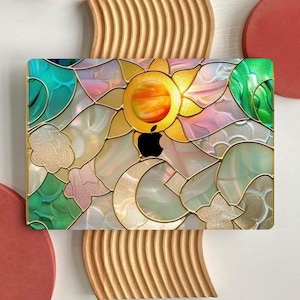 Puede incluir: Una funda de portátil colorida con un diseño de vidriera que representa un sol y elementos florales. La funda tiene un logotipo de Apple negro y está colocada sobre un fondo claro con detalles decorativos de madera y rojos.