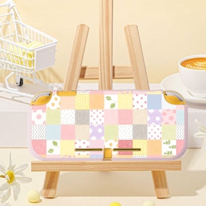 Könnte beinhalten: Eine pastellfarbene Nintendo Switch Lite Hülle mit Patchwork-Design. Die Hülle hat Quadrate in Rosa, Gelb, Blau und Grün mit Blumen- und Tupfenmustern. Die Hülle hat gelbe Akzente und wird auf einer hölzernen Staffelei präsentiert.