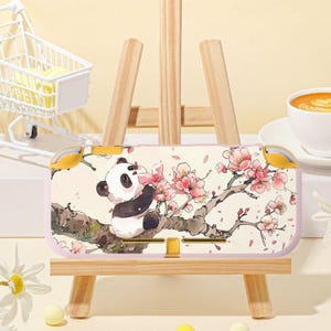 May include: A light pink Nintendo Switch case featuring a panda bear eating a pink flower on a tree branch with pink blossoms. The case is displayed on a small wooden easel. A white shopping cart and a cup of coffee are in the background.