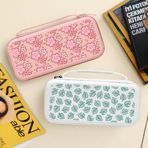 Puede incluir: Dos estuches rectangulares para consola de juegos. Uno es rosa claro con un patrón de flores de cerezo, el otro es blanco con un patrón de hojas verdes. Ambos tienen cierre de cremallera y asa. Sobre una superficie beige claro.