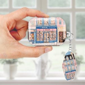 May include: A case and keychain featuring a bookstore design. The case is rectangular with a blue and pink facade, displaying the words "BOOK SHOP". The keychain mirrors the design, with a similar bookstore illustration.